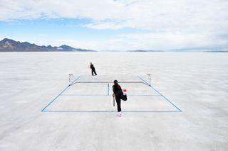 two individuals playing tennis on a salt flat with blue sky and clouds