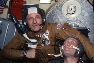 two men in thin helmets with microphones pose for a photograph in front of american and soviet flags inside a spacecraft