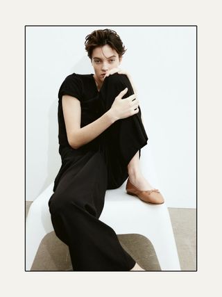 Woman sitting on white chair wearing black T-shirt, black trousers and camel ballet flats