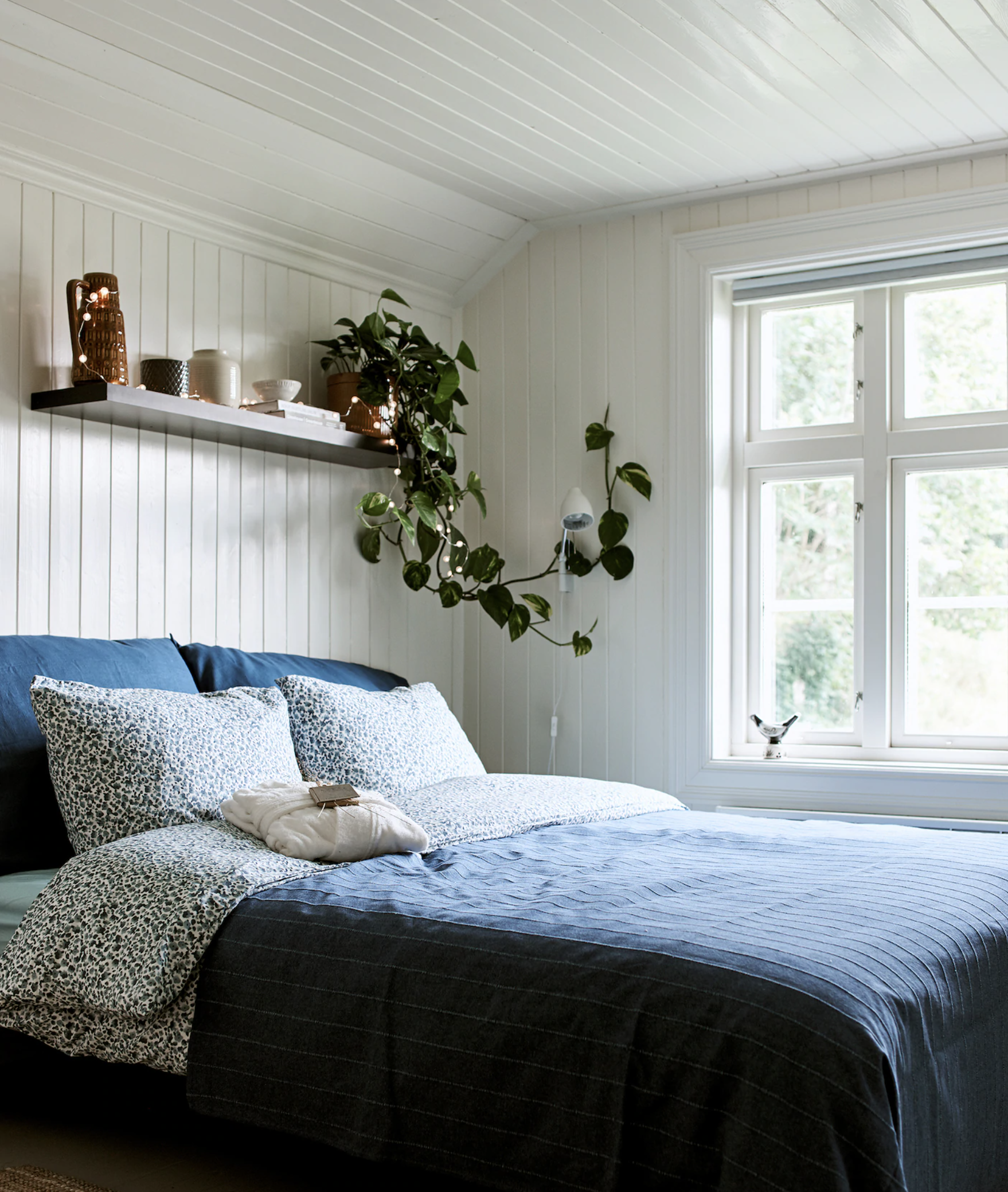 A white bedroom with shiplap on walls and floors, with floating shelves and blue bedding