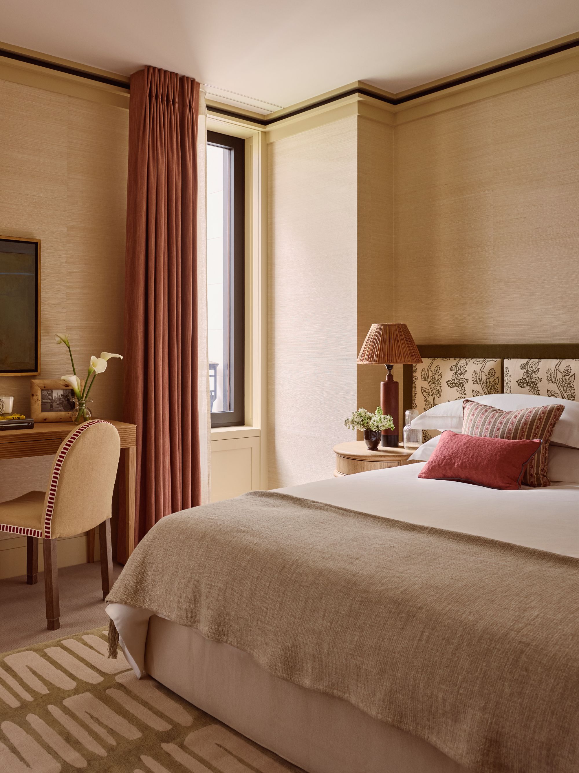 a bedroom with a crisp white bed and red accent pillows, a patterned white and tan carpet, a desk and chair, and floor length curtains blocking a window