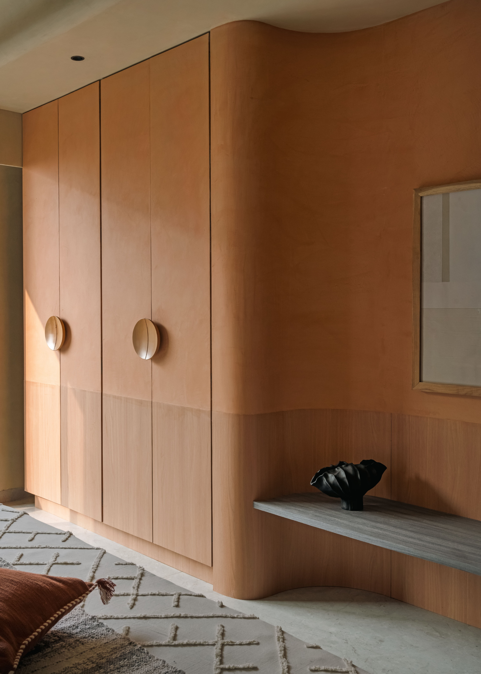A bedroom with a floating bench with a textured ceramic, a hanging painting, a textured rug, an ochre pillow with tassels and a pair of clothes cupboards