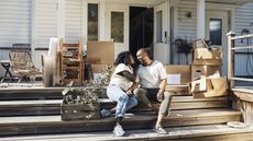 A smiling mature couple sit on their home's front steps, surrounded by moving boxes.