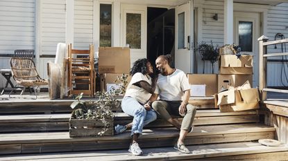A smiling mature couple sit on their home's front steps, surrounded by moving boxes.
