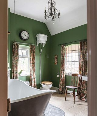 A bathroom with green wall paint decor, long draped curtains, chandelier and khaki bathtub