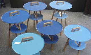 Grey floor, black wall, cluster of metal lid table designs with wooden legs and handles, in various shades of pastel blue