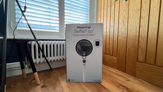 boxed MeacoFan Sefte 10" Pedestal Air Circulator on wooden floor with shuttered window behind, table to left and wooden doors to right