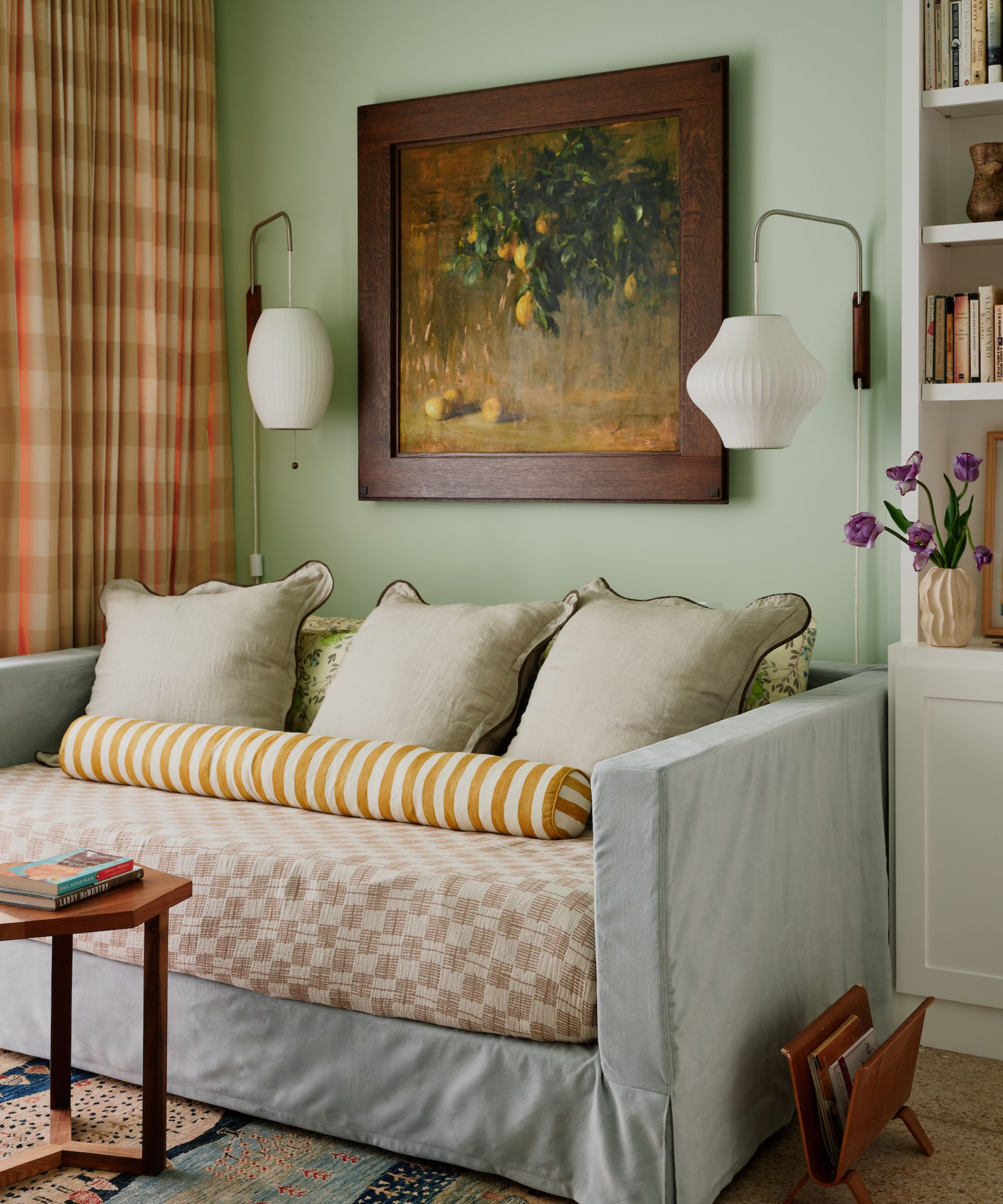 A guest home office with pale green walls, a sofa bed with layered textiles in soft green, ochre, and pink. Checked orange and pink curtains, vintage artwork on the wall, and two modern wall lamps.