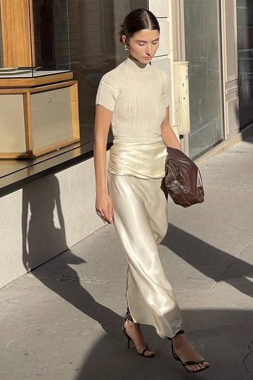 Best strappy sandals shown in a photo of a French woman standing on the sidewalk wearing a white turtleneck short-sleeve top with a white silk maxi skirt and black strappy heel sandals