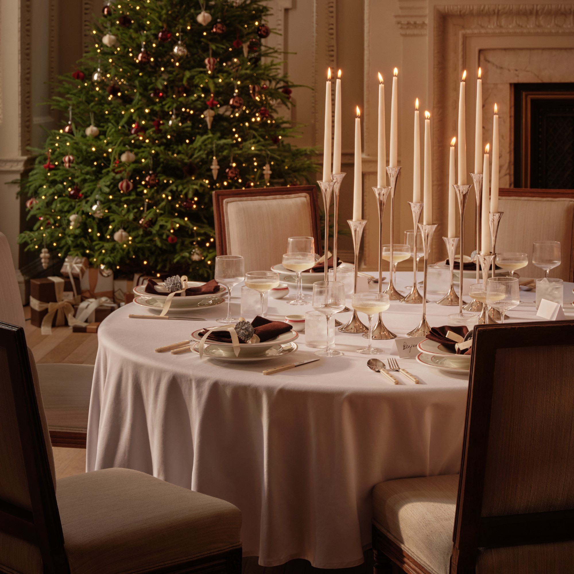 A dining room with a round table with tall candle holders and tapered candles and a decorated and lit-up Christmas tree in the corner
