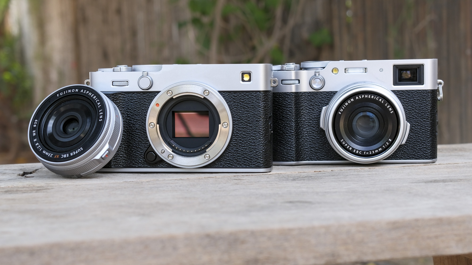 Fujifilm X-E5 alongside the Fujifilm X100VI, on a wooden bench, with the 23mm f/2.8 lens removed from the X-E5