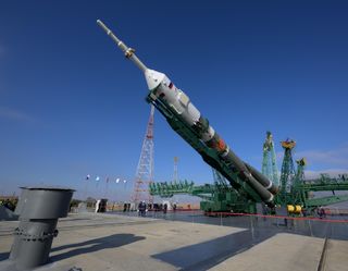 A green/grey soyuz rocket with a white pointy top is laying diagonally as a green support arm that stretches the rocket's height lifts the vehicle vertical.