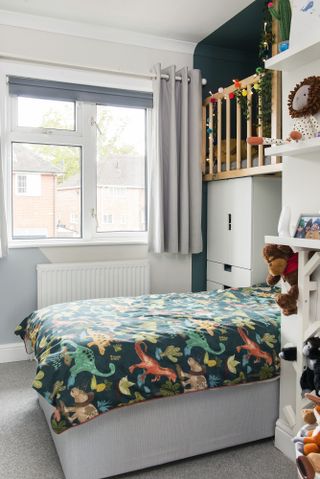 Kid's room with bed covered with dinosaur bedding tucked into an alcove with a den, accessible via a climbing wall, above – painted in a dark green colour with wood railings