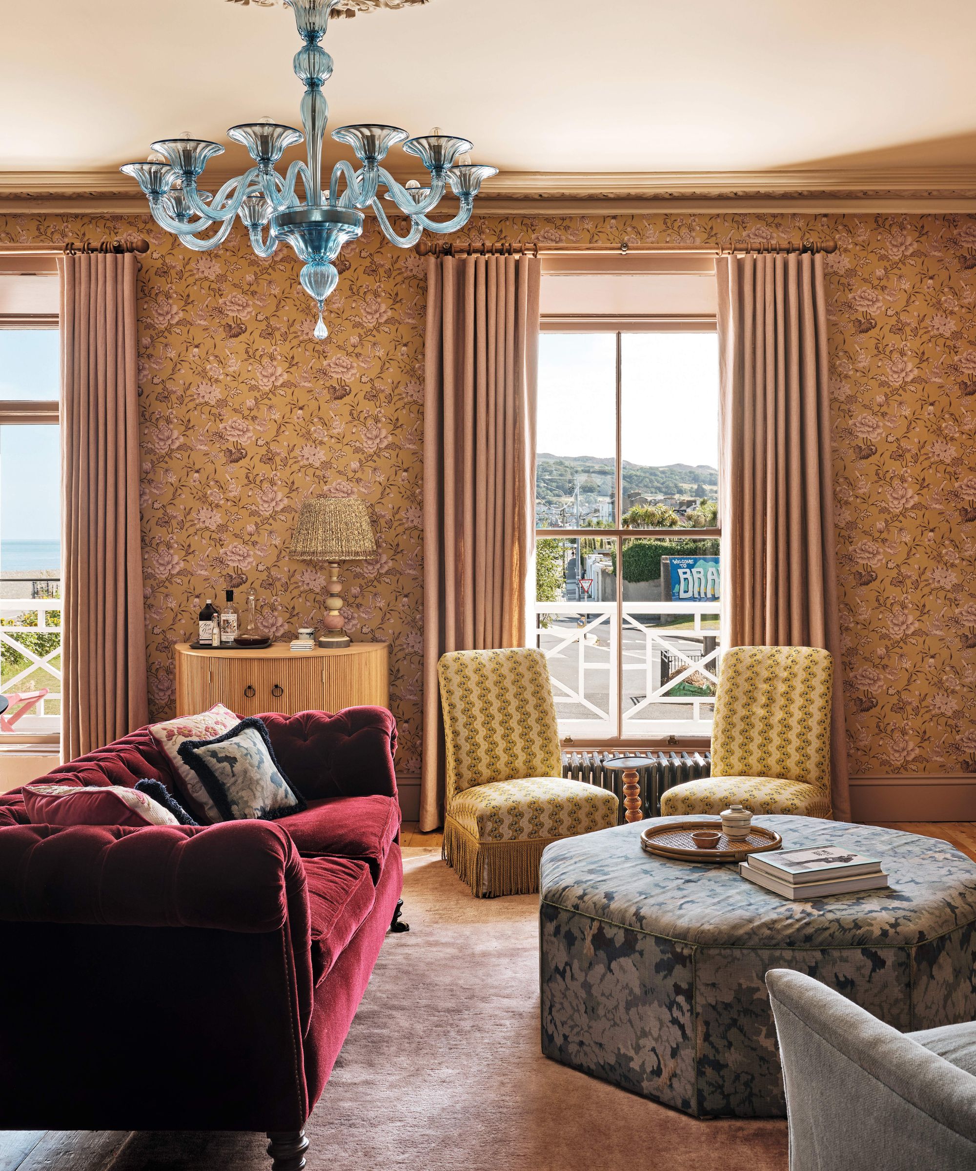 a sunlit sitting room with peach toned floral wallpaper, burgundy velvet couch, yellow slipper chairs, tapestry ottoman and a blue murano chandelier