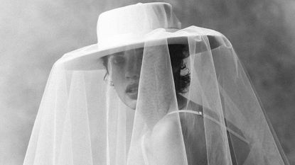 Black and white photo of woman wearing white bra, kneeling down, wearing oversize white Kyha Studios wide-brim bridal hat with veil.