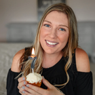 a headshot photo of professional organizer Grace Hall holding a frosted vanilla cupcake with a '7' candle on it: a lady with long blond hair in a black shirt holding a celebratory cupcake looking into the camera indoors