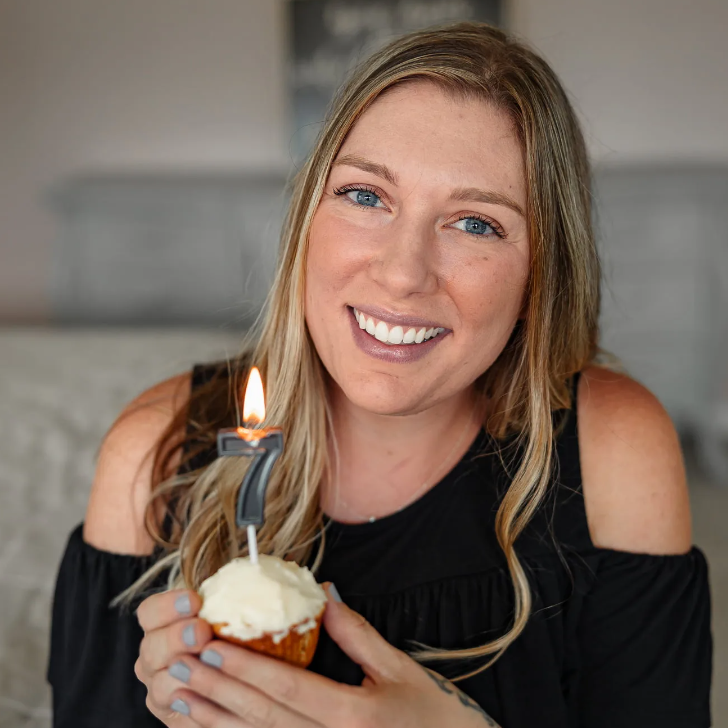 a headshot photo of professional organizer Grace Hall holding a frosted vanilla cupcake with a '7' candle on it: a lady with long blond hair in a black shirt holding a celebratory cupcake looking into the camera indoors