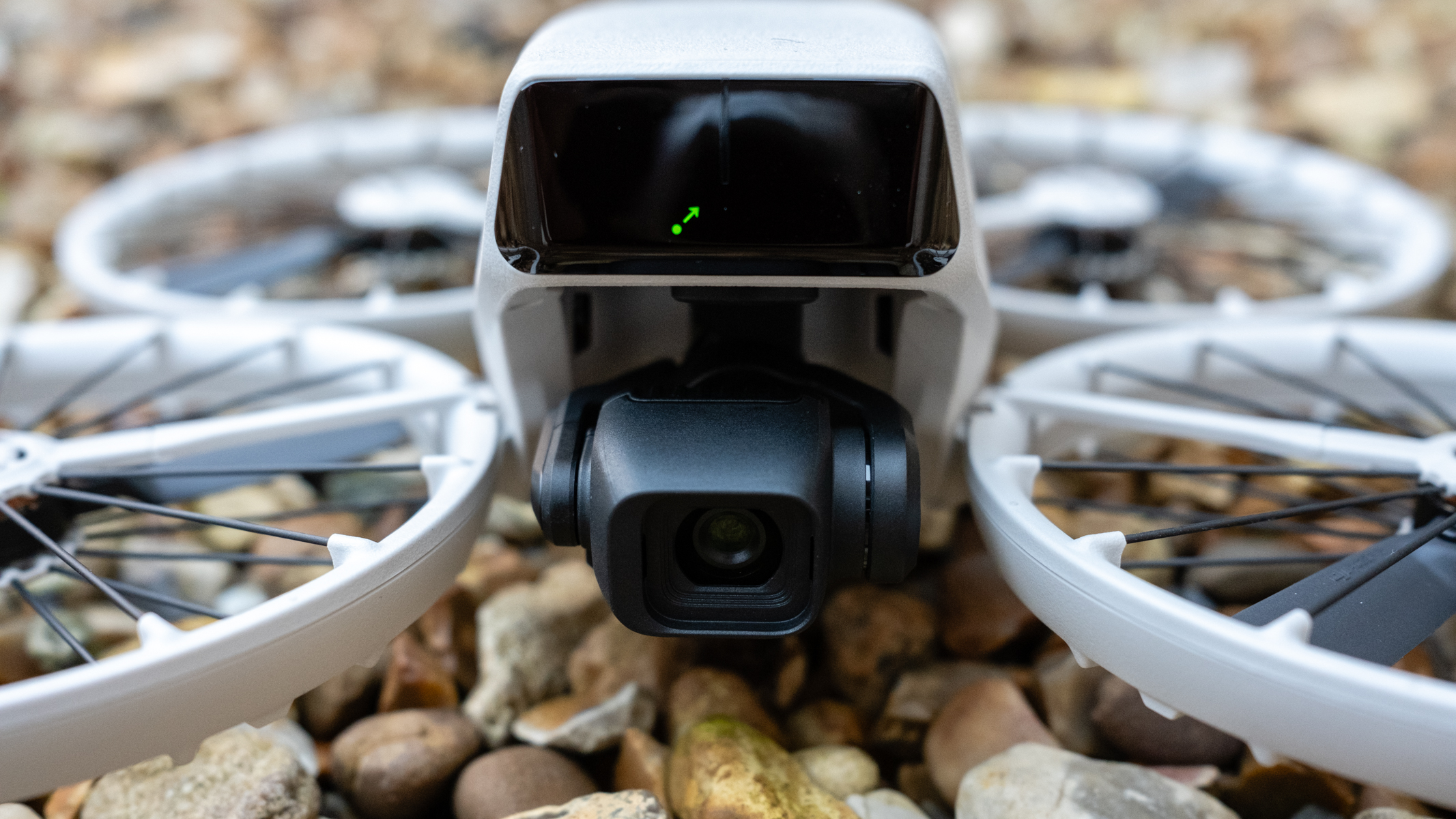 A drone is sitting on a stoney floor, we're looking at it from front on. A black camera is front and centre while four white propellers sit to the side of the white body.