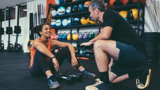 Woman sitting on gym floor with dumbbells between her legs, a man is kneeling and talking to her