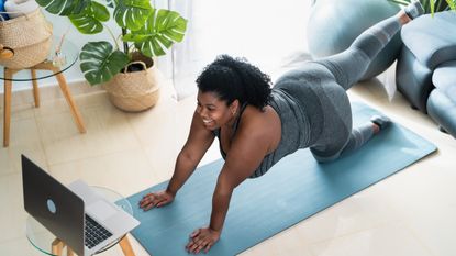 A woman exercises at home on a mat. She is on her hands and knees, with one leg raised in the air behind her. In front of her is an open laptop on a stool and behind her is a couch and plant.