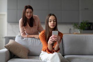 Parental monitoring of teens. Curious mother spying daughter, girl messaging on smartphone on sofa at home. Family and modern technologies