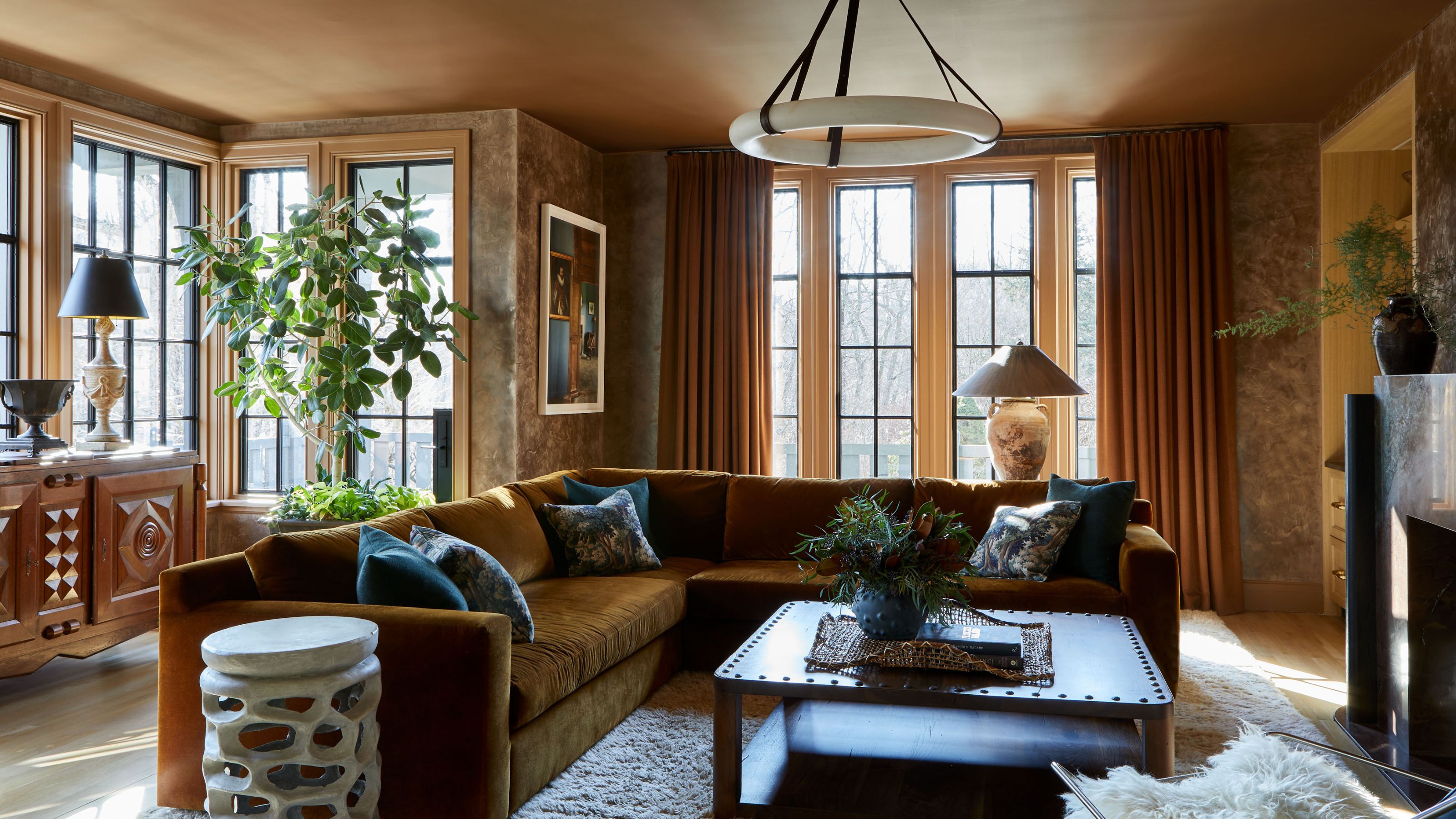 A family room with a cinnamon color palette, with plaster walls and a painted ceiling. A large khaki sectional, a square coffee table with a rug underneath, and a bay window with a large wooden dresser.