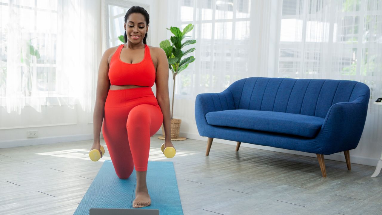 woman in red crop top and leggings performing a reverse lunge on a blue exercise mat with a blue sofa behind her and holding two small yellow dumbbells. 