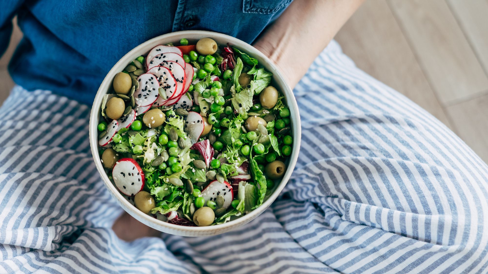 Fibremaxxing: A woman sat cross legged eating a bowl of vegetables, including peas, radish, olives, and salad leaves