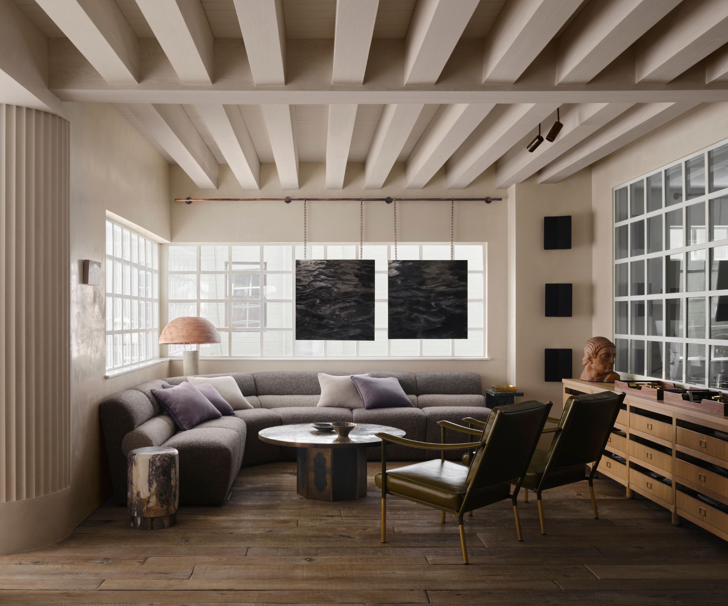 minimalist living room with beige walls and muted purple corner sofa, large horizontal windows, wooden floors, and exposed ceiling beams painted a warm neutral