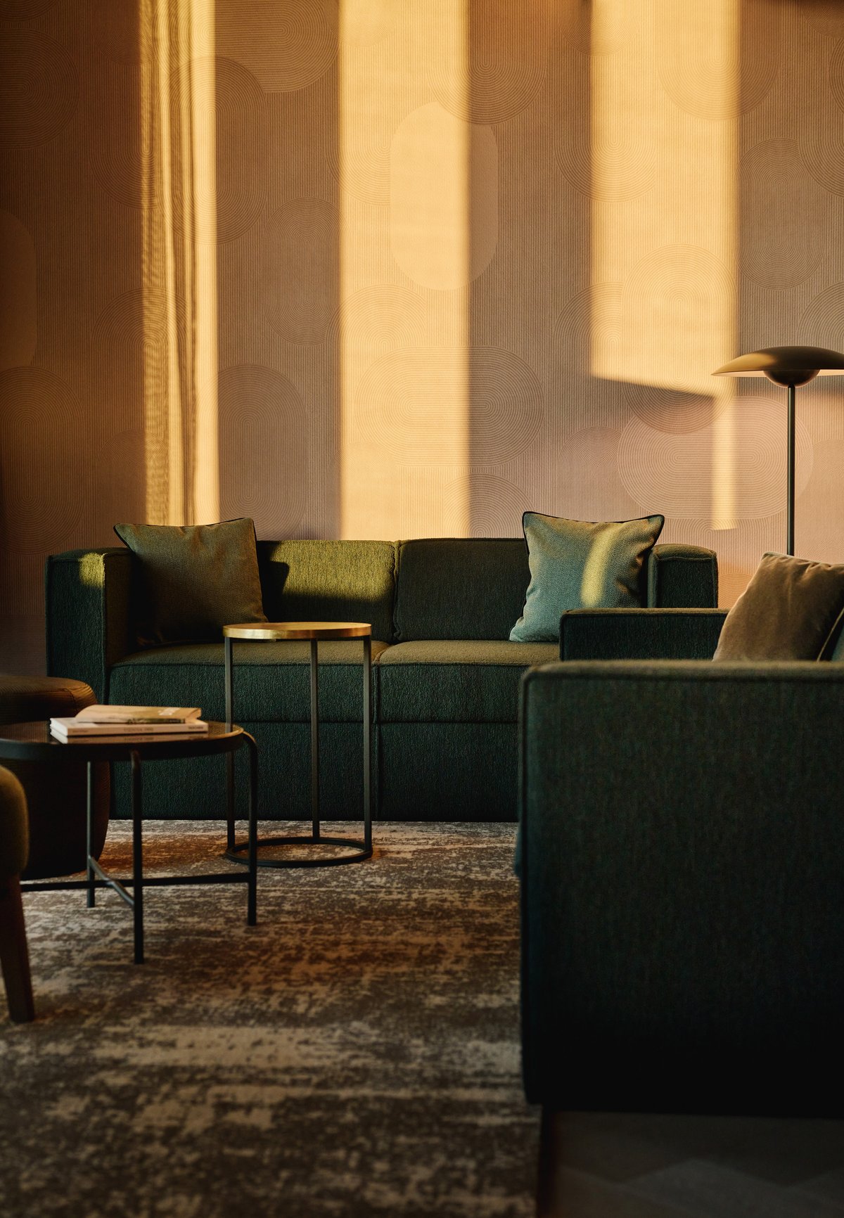 A sun-filled contemporary hotel boasts floor-to-ceiling windows, soft wooden tones mixed with earthy or green shades, mid-century modern-style furniture, and colorful collectibles all around.