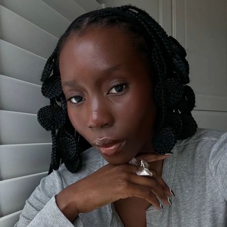 A woman taking a selfie next to a window wearing a grey top with glowing skin
