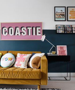 Two-tone wall with dark lower section and light contrast upper half, with mustard velvet sofa, and large pink typo framed wall print.