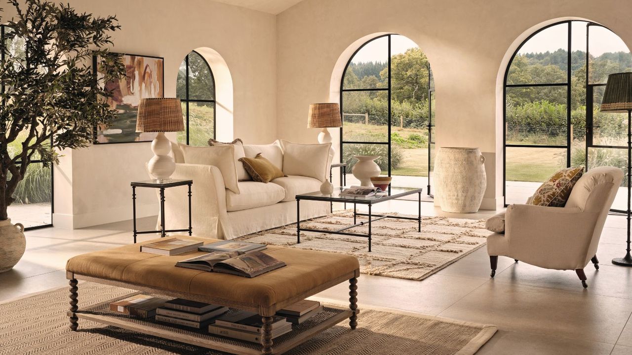 A bright living room with a cream sofa, huge arched windows and a large footstool