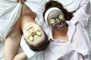 A couple laying next to each other wearing facemasks and slices of cucumbers over their eyes.