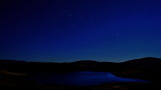 silhouetted hills roll across the landscape with a lake in the foreground and a starry sky in the background.