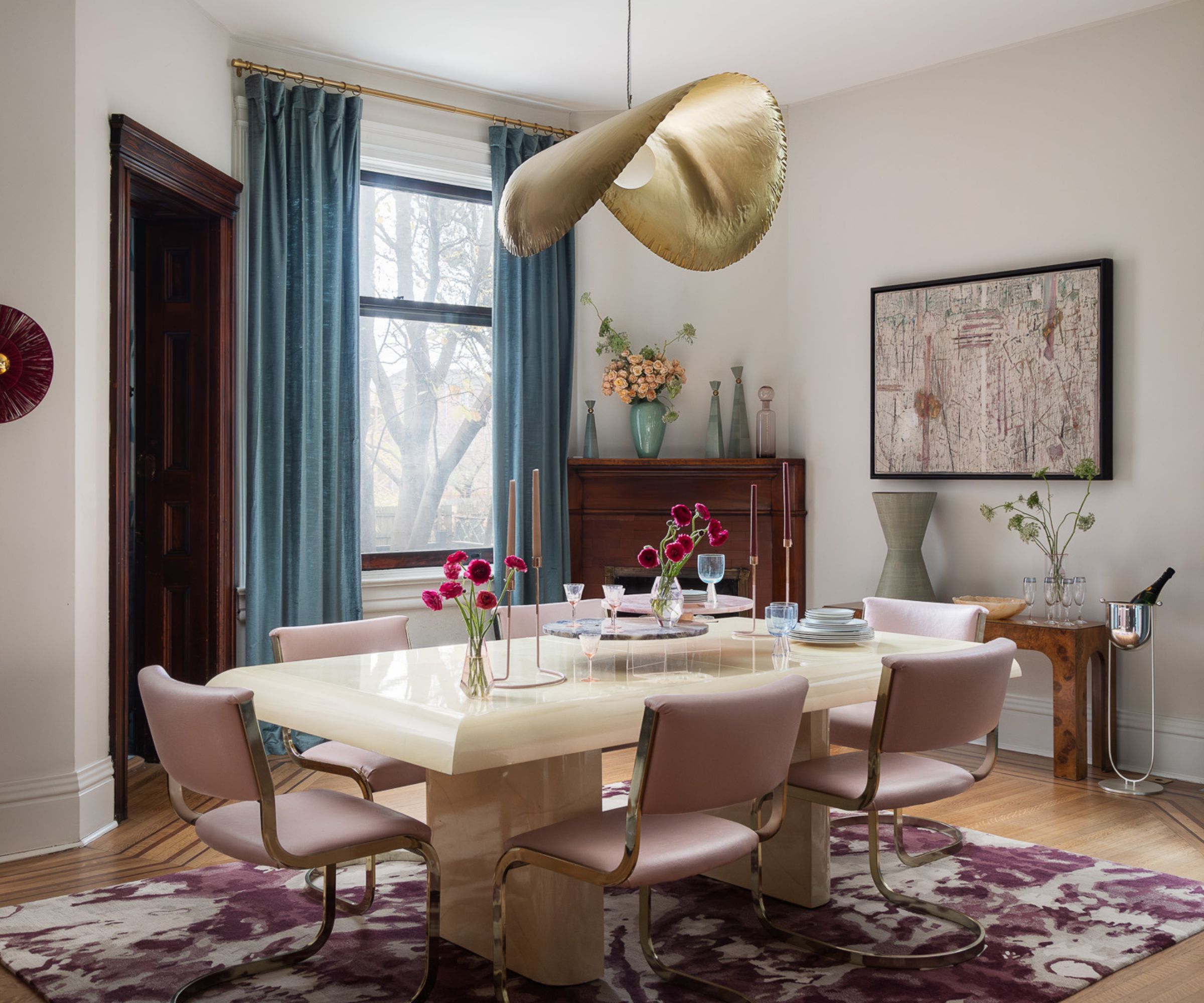 A dining room with eight walls painted white, a rectangular dining table surrounded by six pink chairs, and a statement gold pendant light above