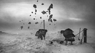 Two people dressed in extreme-cold clothing shovel snow from a frozen surface. 