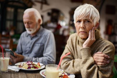 Senior woman feeling depressed while having lunch with her husband who is using cell phone in a restaurant.