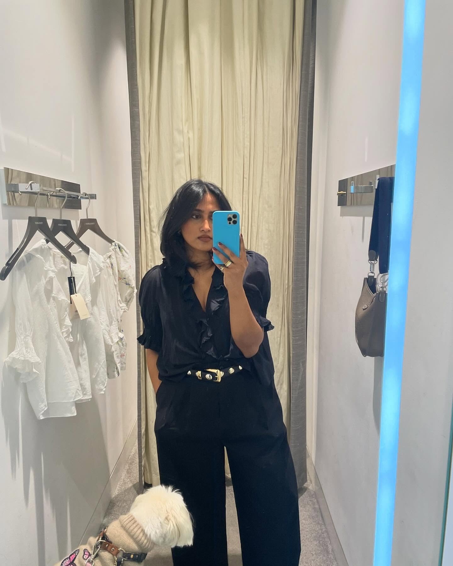 Influencer takes a mirror selfie in a changing room. She's wearing a black frilly blouse tucked into black jeans with a Khaite studded belt looped through her jeans.