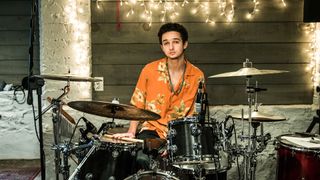 A drummer in an orange shirt waits to play, with his sticks on a drum
