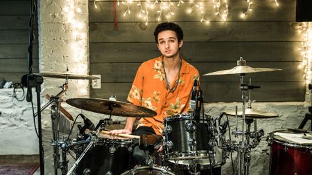 A drummer in an orange shirt waits to play, with his sticks on a drum