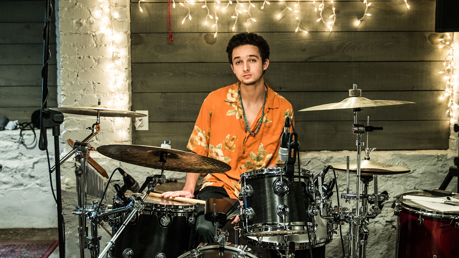 A drummer in an orange shirt waits to play, with his sticks on a drum