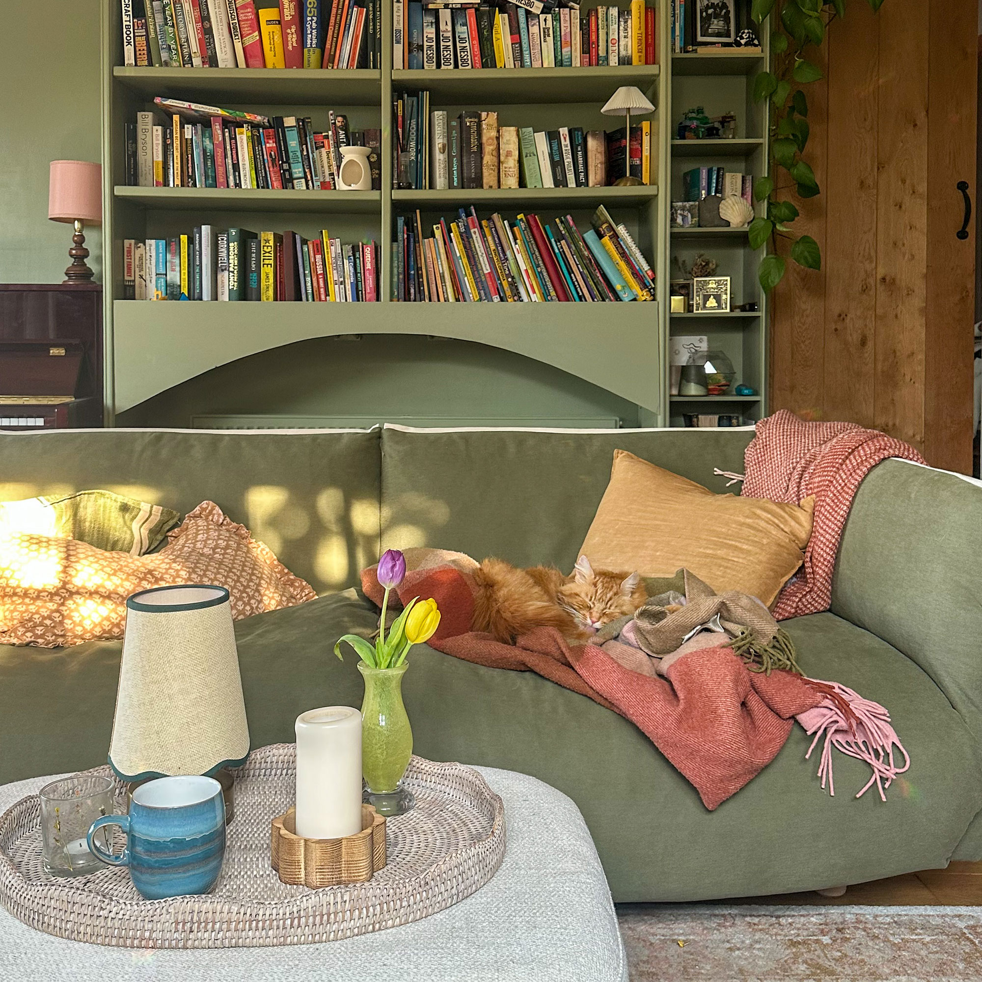 Green living room with built-in shelving and green sofa in front cream fabric ottoman as coffee table with tray holding a mug, lamp, candle and vase