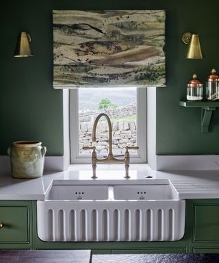 Green painted kitchen with artistic, mural style blind, deep white sink, golden wall lights either side of blind, window with view of countryside