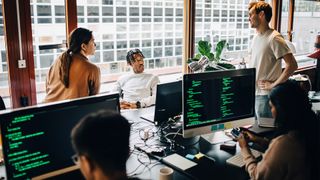 Cybersecurity team members discussing strategy in an open plan office space, with male and female practitioners standing and others sitting at desks.