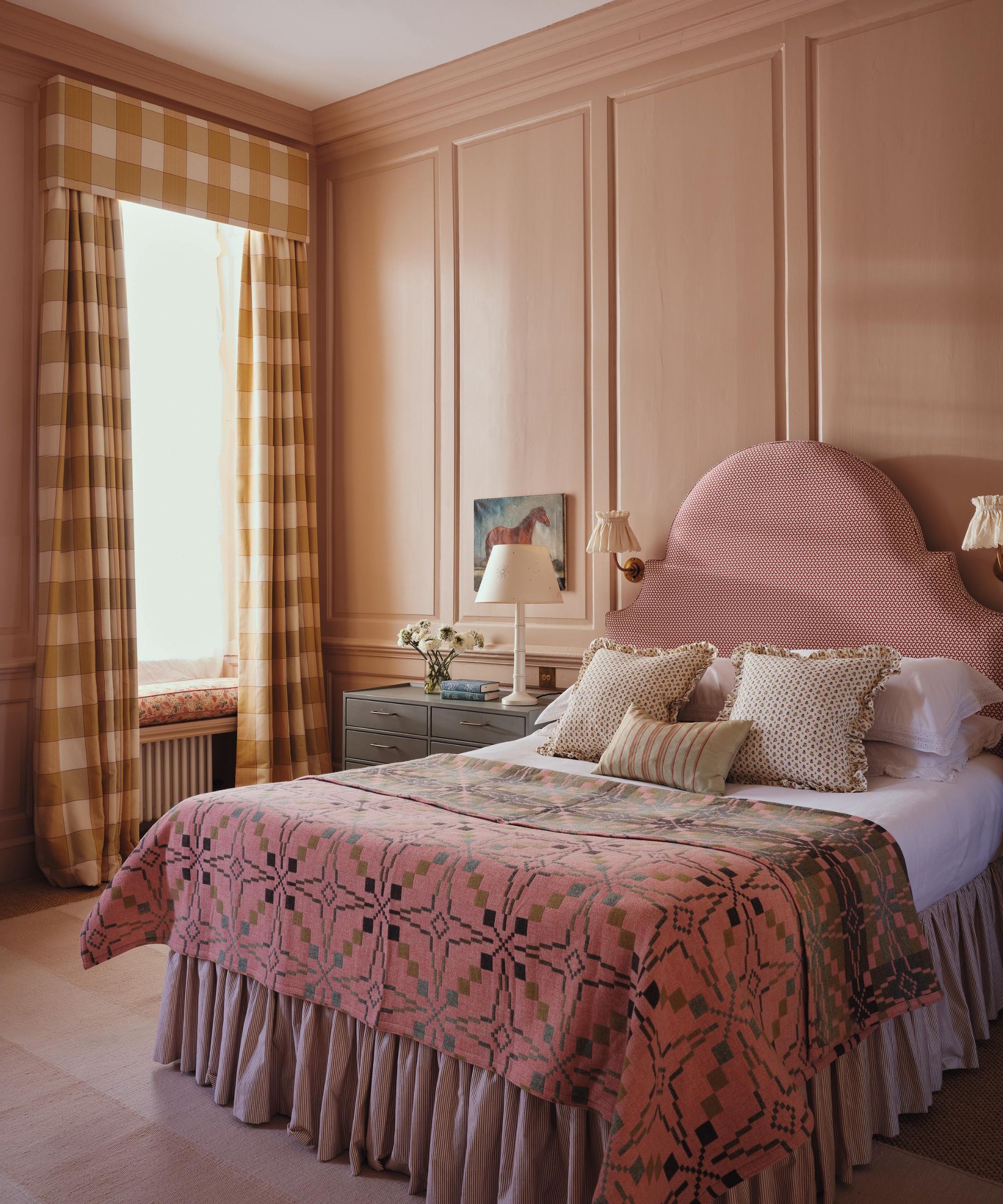 a traditional english pink bedroom with wall moldings, a large Georgian window with yellow gingham window dressings, pink upholstered ed styled with pillows and a Welsh blanket