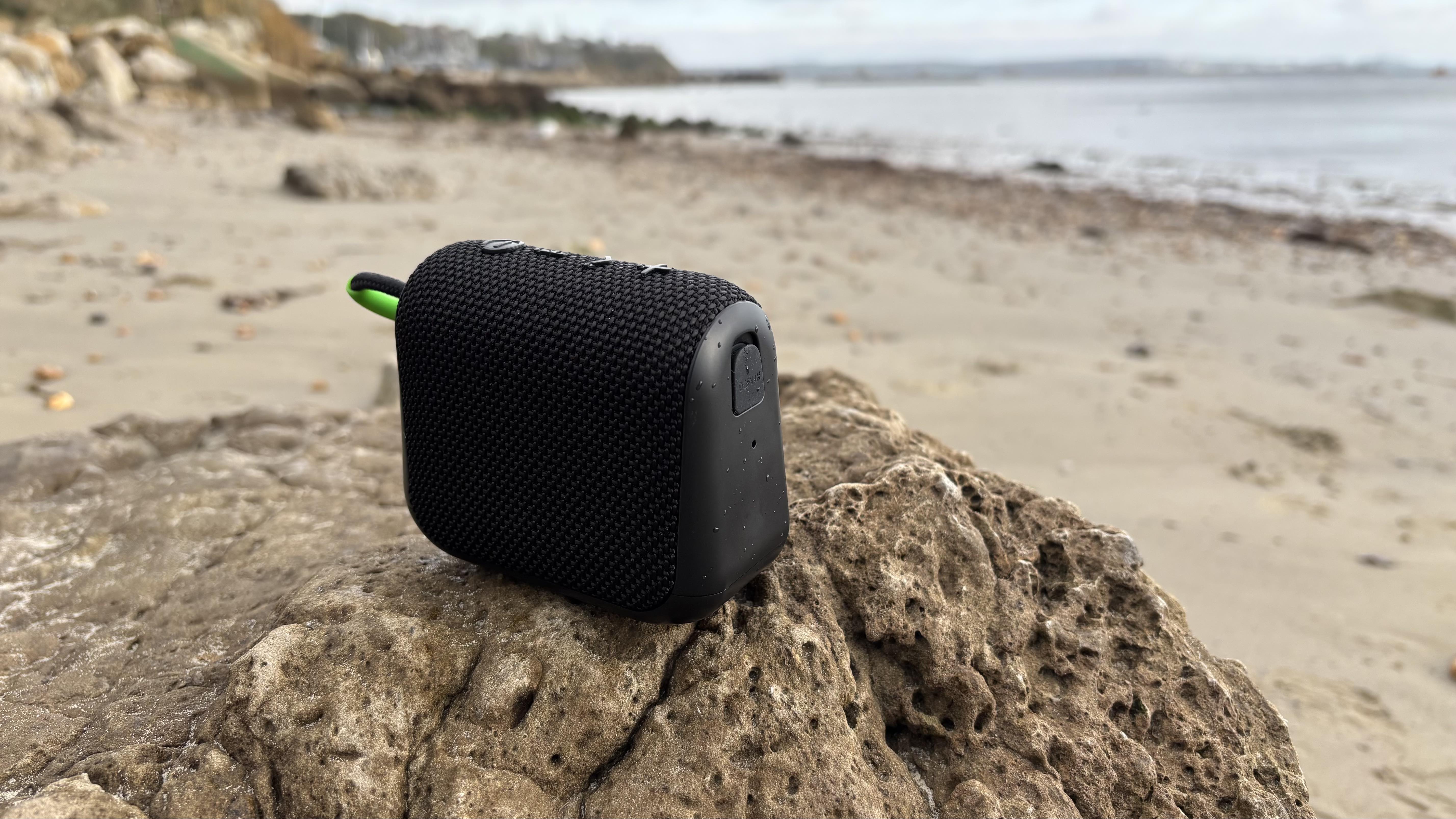 Black AO mini portable wireless speaker on a rock, with a beach and the sea in the background