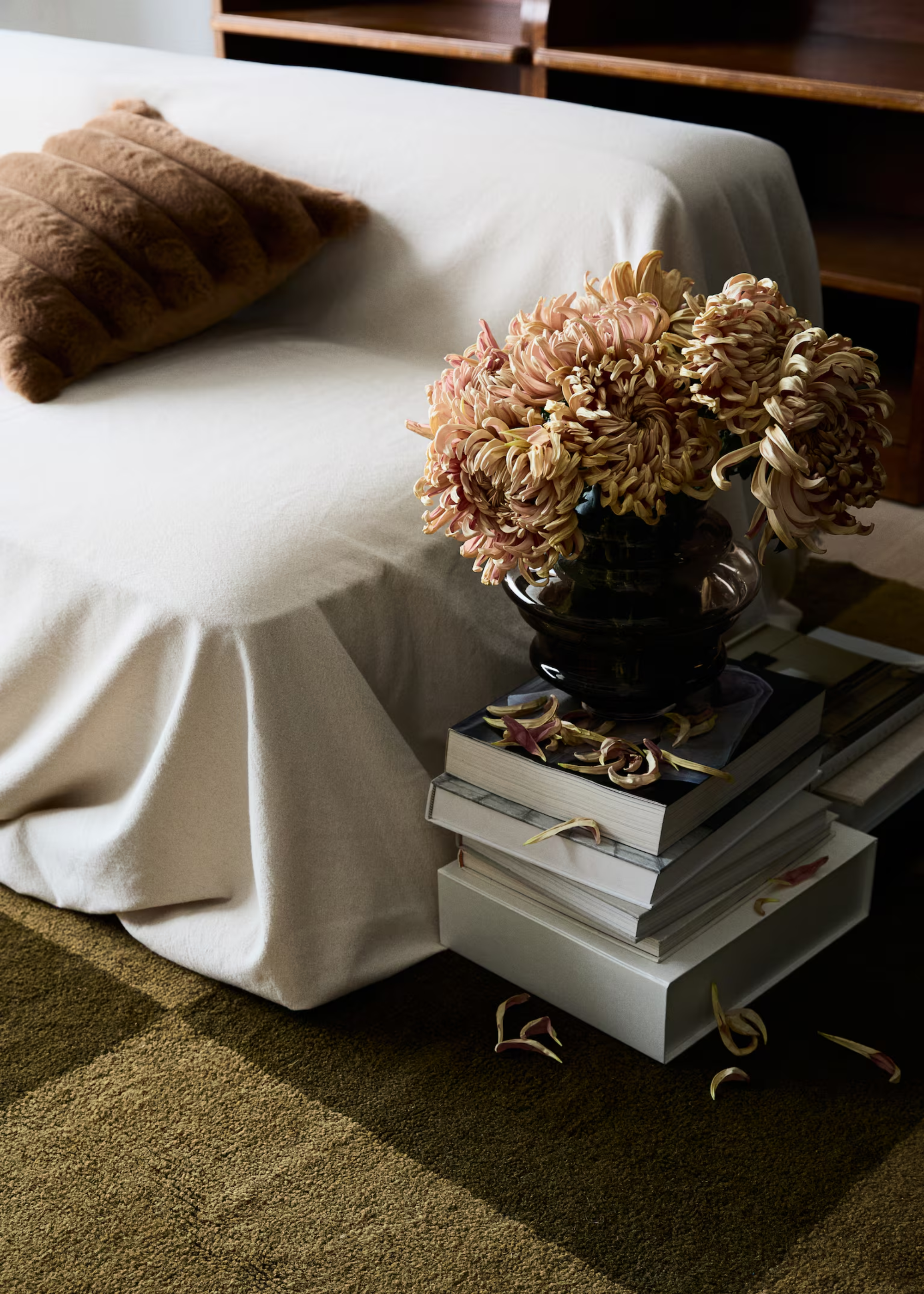 A sofa with a beige sheet over it with a fuzzy brown cushion by a stack of books with a dusty brown glass vase with chrysanthemums