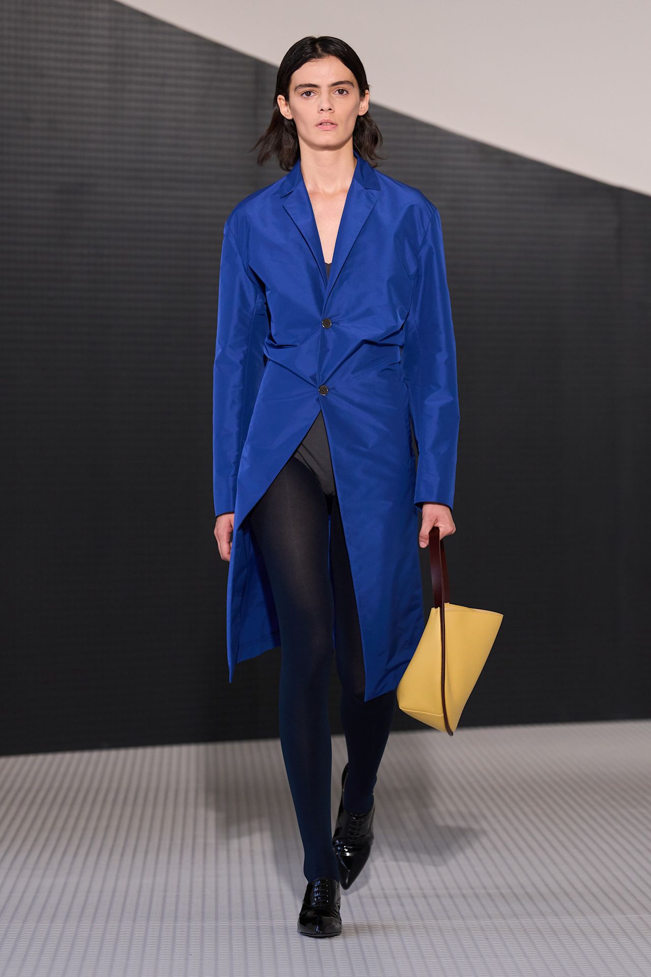 A Jil Sander model wearing a bright blue coat layered over a gray bodysuit, black tights, black pointed derby shoes, and a yellow crescent shoulder bag.