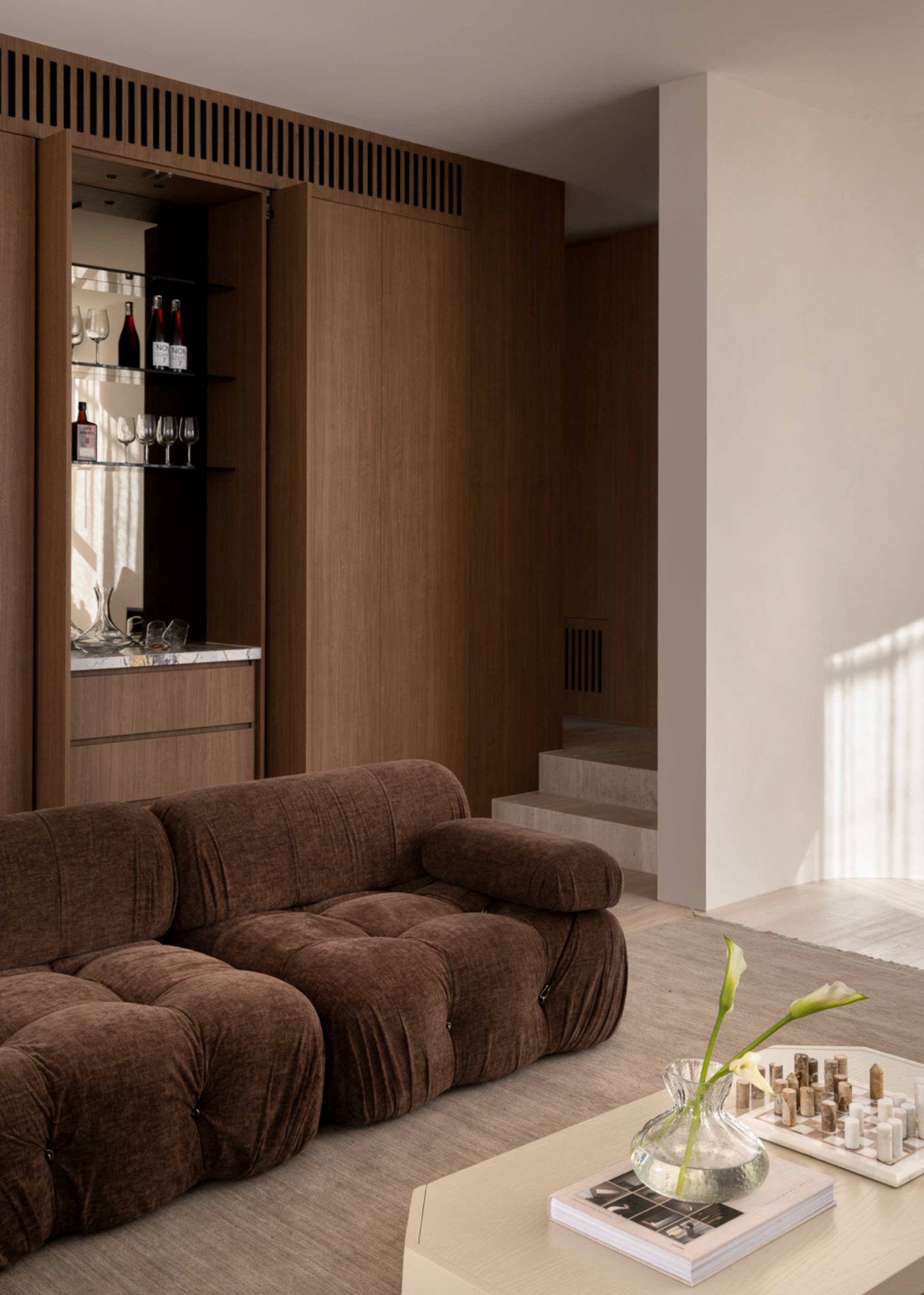 A living room with a home bar nook, a soft brown sofa, a white coffe table with a vase of flowers, a book, and a chess set
